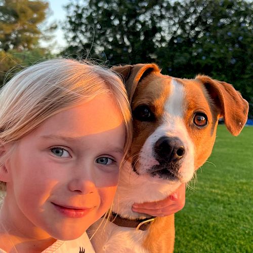 A young girl with blonde hair smiles while holding a brown and white dog on a green lawn with trees in the background under sunny skies.