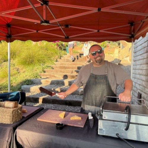 A man smiling under a red canopy tent, preparing food on a grill with a spatula in hand. There are some cut sandwiches on a cutting board.