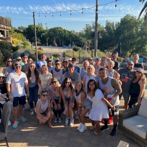 A group of people posing together outdoors, likely at a gathering or party, with some seated and others standing, near a pool and a bounce house.