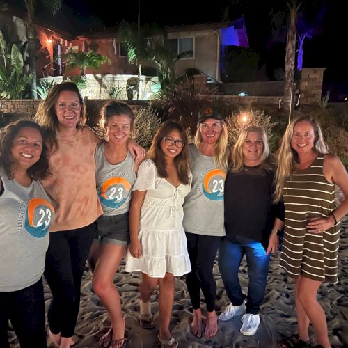 A group of seven people standing together on a sandy beach at night, with smiles and casual clothing.