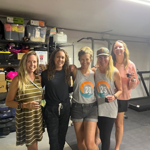 Five women standing in a garage, posing for a photo, smiling, with shelving and storage boxes in the background.