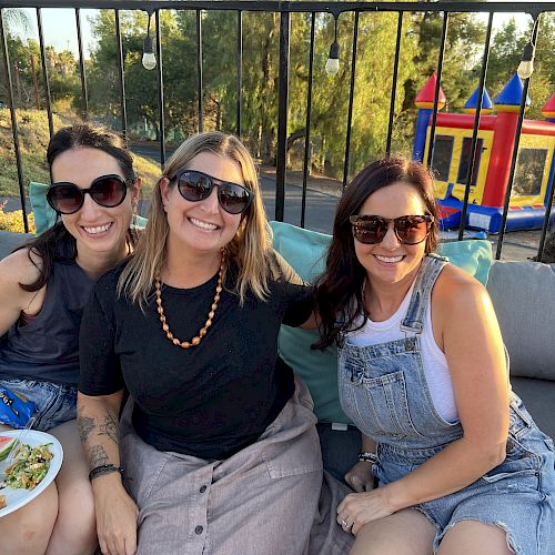 Three women sitting on a sofa outdoors, smiling, wearing sunglasses. There’s a plate of food; a colorful inflatable structure is in the background.