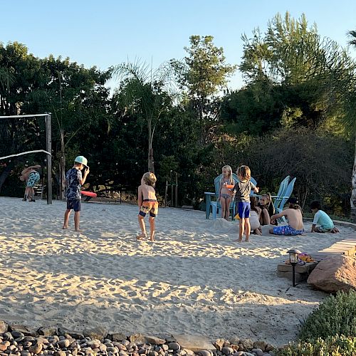 Children are playing on a sand court while adults relax in chairs nearby, surrounded by greenery and clear skies.