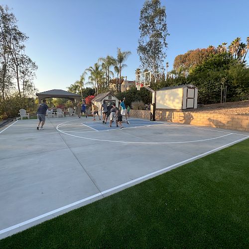 The image shows a group of people playing basketball on an outdoor court surrounded by trees and a well-maintained lawn area.