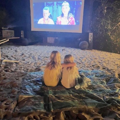 Two people sitting on a blanket, in front of an outdoor movie screen displaying a movie, under a starry night sky.