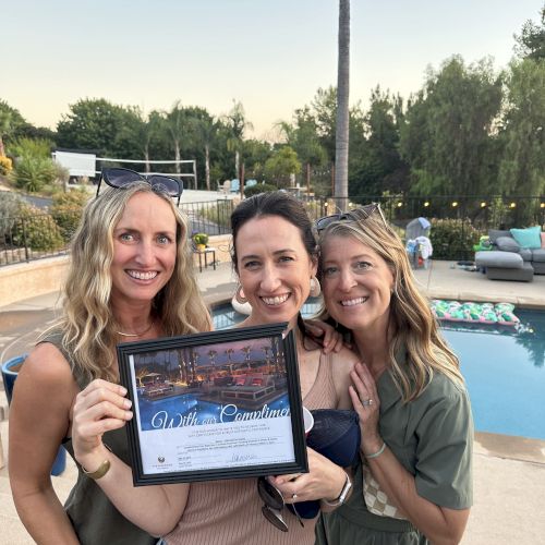 Three people smile by a pool, holding a framed certificate. Sunglasses and outdoor decor are visible in the background.