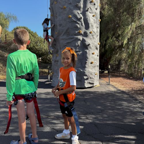 Children in harnesses are near a climbing wall. One kid is climbing while others wait. Background includes trees and clear sky.