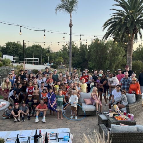 A large group of people gathered outdoors, with string lights, palm trees, and patio seating, enjoying a social event.