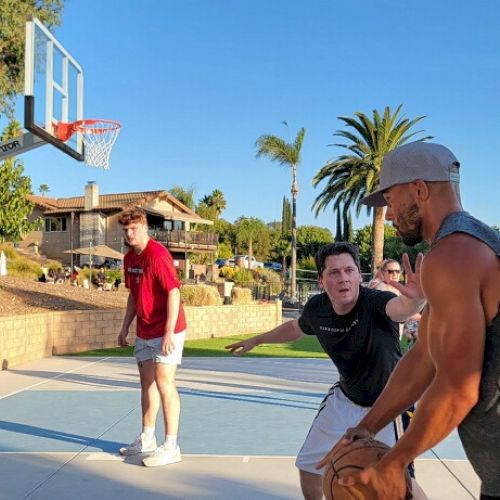 A group of people are playing basketball outdoors on a sunny day, with trees and a house in the background.