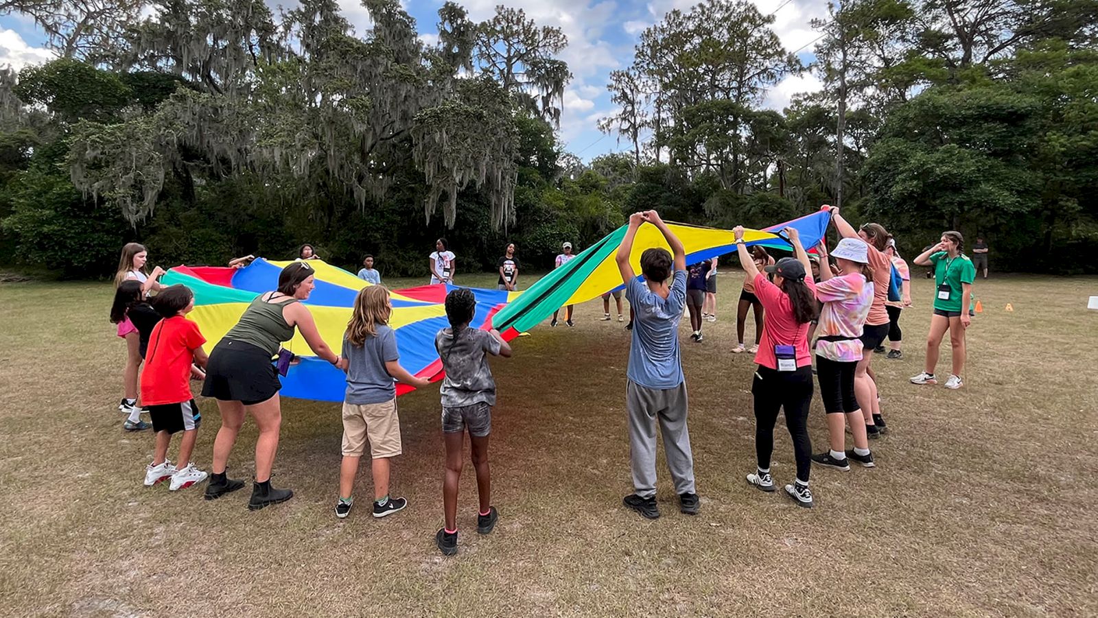 A group of people is outside, holding a colorful parachute in a circle with trees in the background.