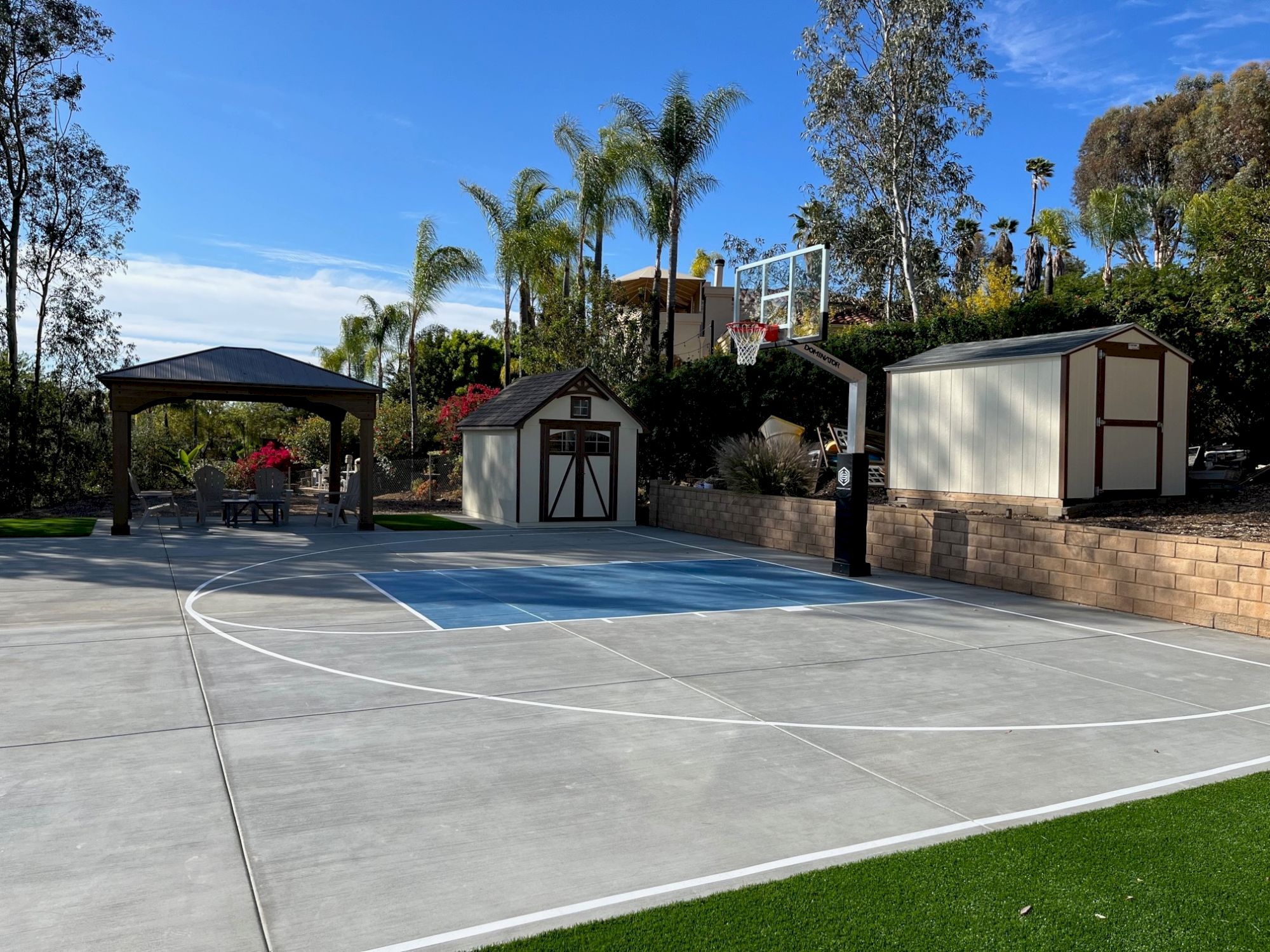 A backyard features a basketball half-court, a gazebo with outdoor seating, a small storage shed, and lush landscaping with tall trees and greenery.