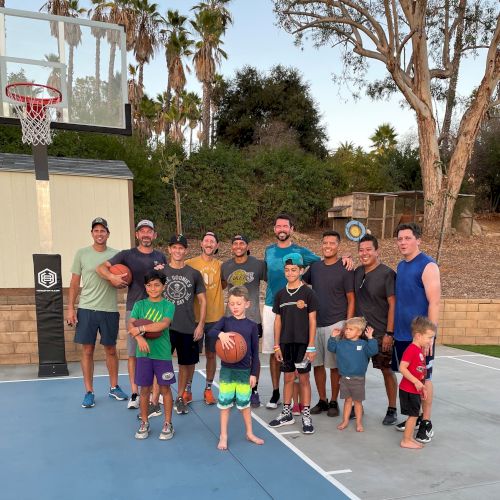 A group of people, including adults and children, are gathered on a basketball court, some holding basketballs. Trees and a shed are visible in the background.