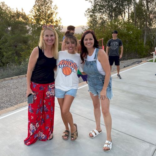 Three women are posing and smiling outdoors on a paved area; two people are in the background among trees. Always ending the sentence.